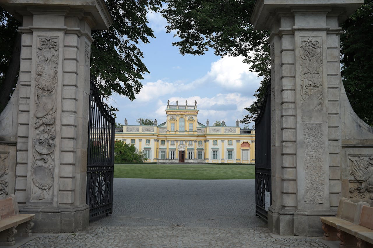 View of a historic palace through an ornate gate with decorative pillars on a sunny day.
